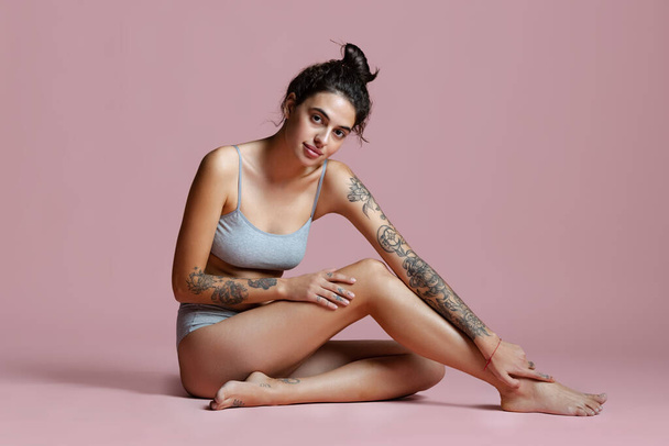 Full-length portrait of beautiful young woman sitting in gray cotton underwear isolated over pink background. - Fotografie, Obrázek