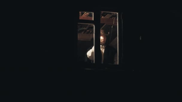 Ένας άντρας με καουμπόικο καπέλο στη σοφίτα του σπιτιού. Δίνει σήμα με κερί από το παράθυρο. - Πλάνα, βίντεο