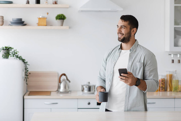 Smiling middle aged caucasian male with beard with smartphone reading news and drinking coffee - Photo, image