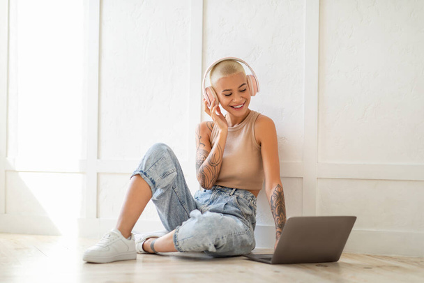 Junge Dame in drahtlosen Kopfhörern mit Laptop, auf dem Boden über Lichtwand sitzend, Musik hörend und auf der Tastatur tippend - Foto, Bild