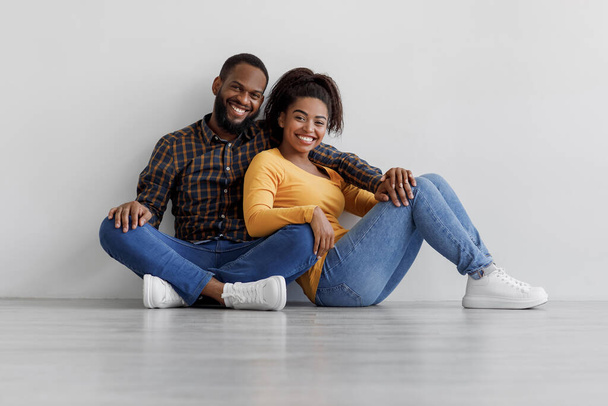 Happy millennial african american couple hugging sits on floor in empty room on white wall background, free space - Foto, afbeelding