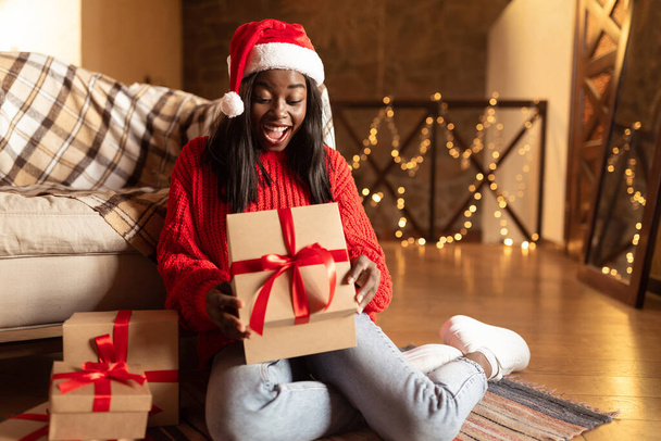 Święta i Nowy Rok. Podekscytowany młoda czarna kobieta w Santa kapelusz otwarcie prezenty świąteczne na podłodze w domu - Zdjęcie, obraz