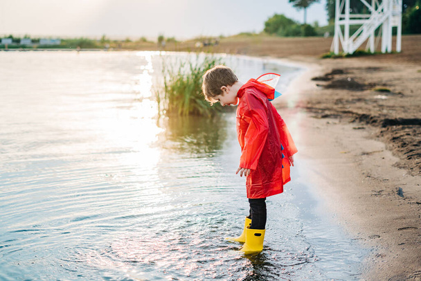 Junge in rotem Regenmantel und gelben Gummistiefeln beim Wasserspielen am Strand. Schulkind im wasserdichten Mantel springt im Wasser auf See. Kind hat Spaß mit Wellen am Ufer - Foto, Bild