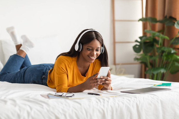 Mujer negra contenta usando teléfono usando auriculares inalámbricos en casa - Foto, imagen
