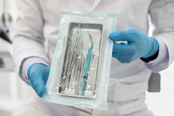 Γιατρός φορώντας μπλε αποστειρωμένα γάντια επιδεικνύοντας σφραγισμένη θήκη αποστείρωσης με οδοντικά εργαλεία - Φωτογραφία, εικόνα