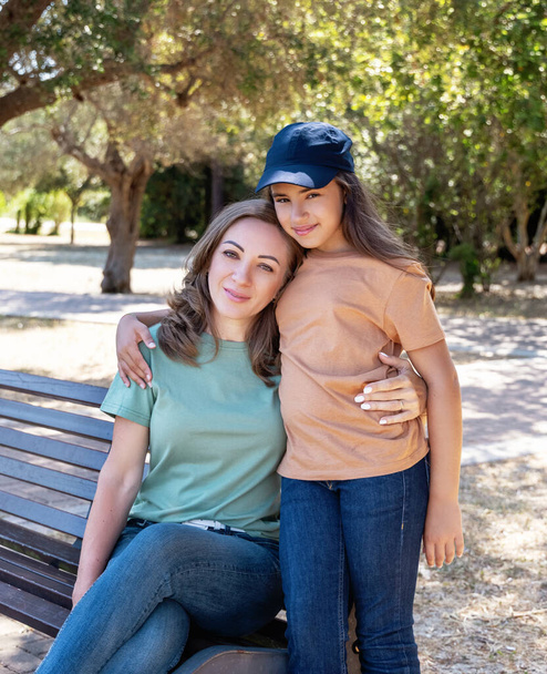 Mother and daughter sitting on the wooden bench in the park in sunny day. A middle-aged woman and kid girl wearing shirts and jeans. T-shirts mockup. Family spending time together in summer - Photo, Image