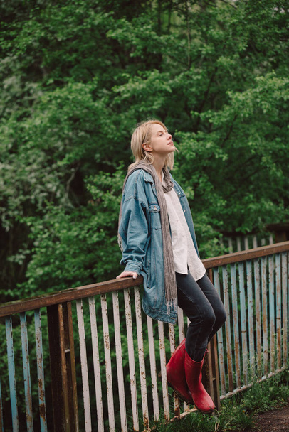 Blonde in demin jacket and red boots on a bridge near river at country - Фото, зображення