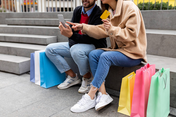 Cropped view of young couple with shopper bags, smartphone and credit card sitting on stairs outdoors, shopping online - Foto, imagen