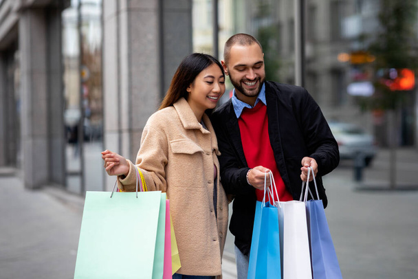 Liebevolles multinationales Paar steht in der Nähe des Supermarktes, schaut in Einkaufstüten, geht gemeinsam einkaufen - Foto, Bild