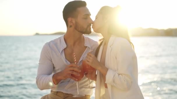 Χαμογελαστό ζευγάρι ισπανόφωνων με κοκτέιλ που φιλιούνται στη θάλασσα - Πλάνα, βίντεο