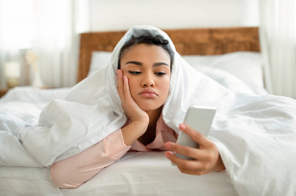 Rozrušená mladá černoška se dívá na obrazovku smartphonu, zatímco leží v posteli pod dekou, cítí se osamělá nebo deprimovaná - Fotografie, Obrázek
