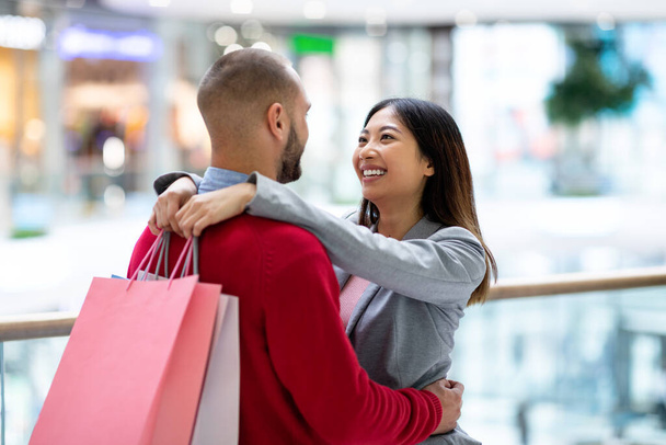 Schönes junges, vielseitiges Paar mit Einkaufstüten, die sich in einem großen Supermarkt umarmen und anschauen - Foto, Bild