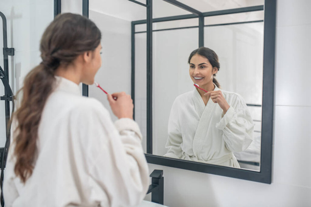 A young pretty woman in a white bathrobe brushing her teeth - Foto, imagen