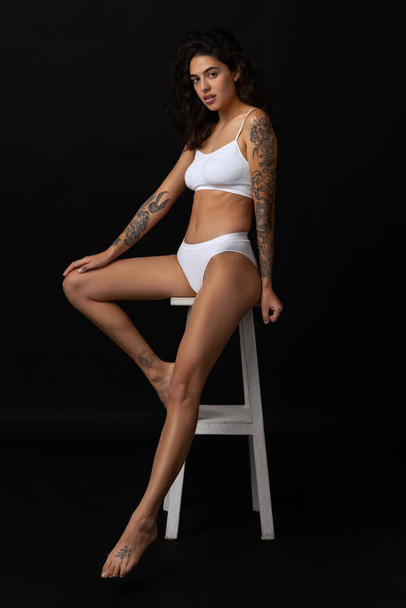 Full-length portrait of beautiful slim girl in white cotton underwear posing on chair isolated over dark background - Photo, Image