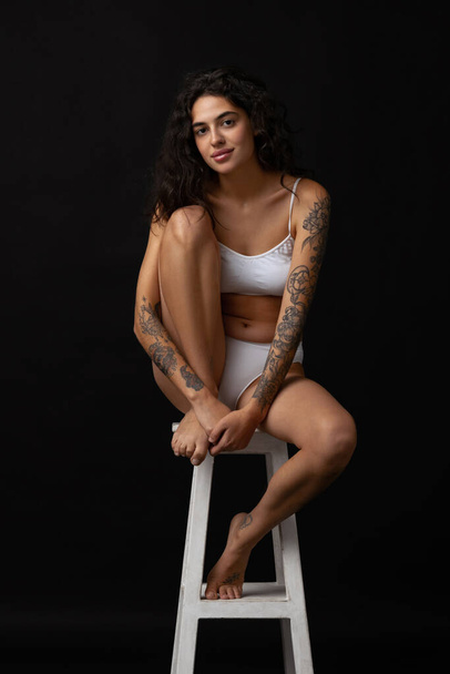 Full-length portrait of beautiful slim girl in white cotton underwear posing on chair isolated over dark background - Foto, Bild