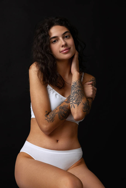 Cropped portrait of beautiful slim girl in white cotton underwear posing on chair isolated over dark background - Φωτογραφία, εικόνα