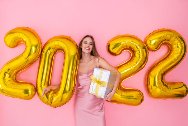 Smiling girl stands near 2022 air balloons holds gift box on pink background. New Year celebration - Foto, Imagen