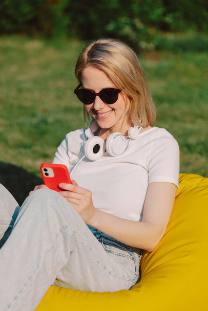 Loira em óculos de sol e fones de ouvido com telefone celular sentado em saco de feijão amarelo em um jardim - Foto, Imagem