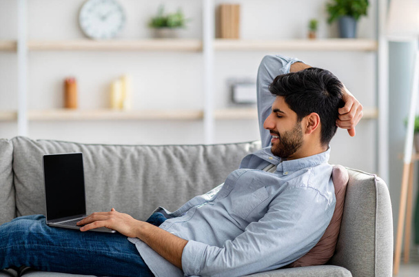 自宅のラップトップコンピュータとソファに座っている若いアラブ人男性、空白のPC画面上のウェブサイトのためのモックアップ。モックアップ - 写真・画像