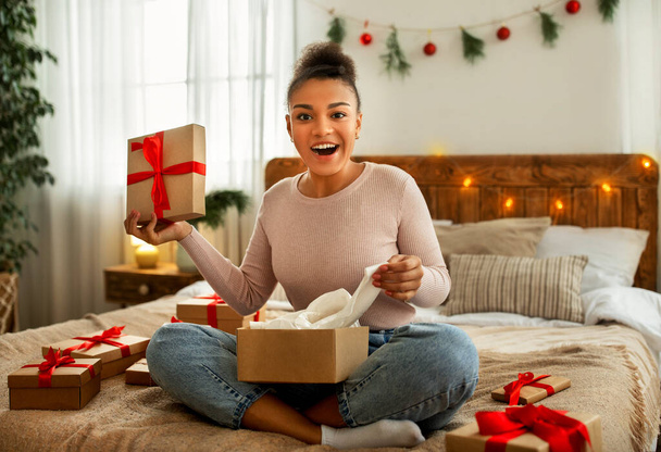 Retrato de señora negra que recibe regalos de Navidad y desempaqueta la caja de cartón, sentada en la cama en el dormitorio acogedor - Foto, Imagen