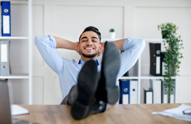 Ευτυχισμένη χιλιετή Άραβας χαλαρώνοντας μετά από επιτυχημένη δουλειά, βάζοντας τα πόδια στο τραπέζι στο σύγχρονο γραφείο - Φωτογραφία, εικόνα