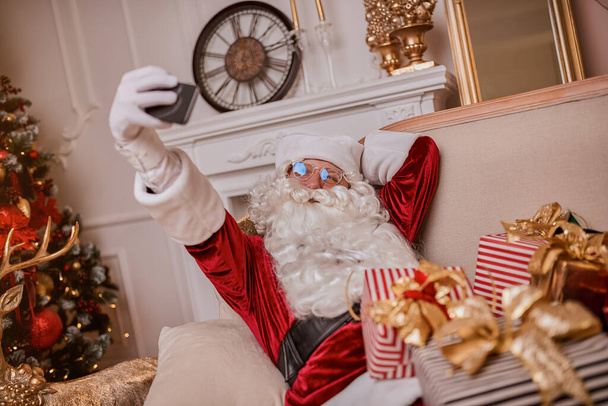 Άγιος Βασίλης κάθεται στον καναπέ και μιλάει στο κινητό τηλέφωνο κοντά στο τζάκι και χριστουγεννιάτικο δέντρο με δώρα. Νέο έτος και Καλά Χριστούγεννα, καλές γιορτές έννοια - Φωτογραφία, εικόνα