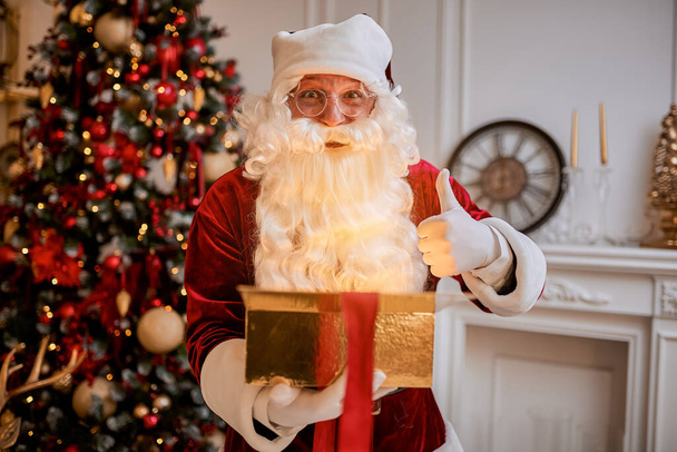 Surprised Santa Claus with a magical glowing gift near beautiful christmas tree.  New year and Merry Christmas , happy holidays concept - Photo, Image