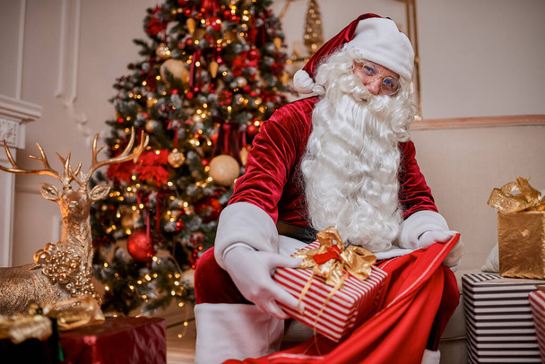 Święty Mikołaj wkłada prezenty do swojej wielkiej czerwonej torby, aby życzyć dzieciom Wesołych Świąt. - Zdjęcie, obraz