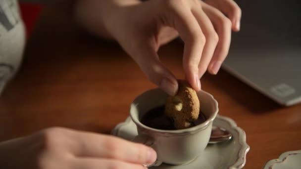 4k gros plan de la main féminine biscuit trempette à la main dans une tasse de café aromatique. Vintage tasse à café est placé sur une table en bois près d'un ordinateur portable, concept de pause café et petit déjeuner lent, mise au point douce - Séquence, vidéo