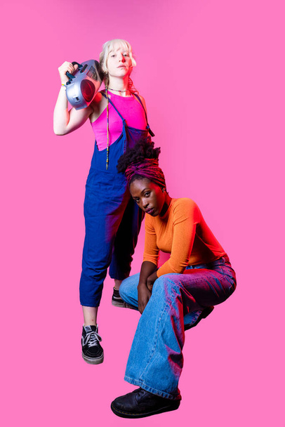 Две молодые разноплановые женщины держат бумбокс, позируя уверенно - Фото, изображение