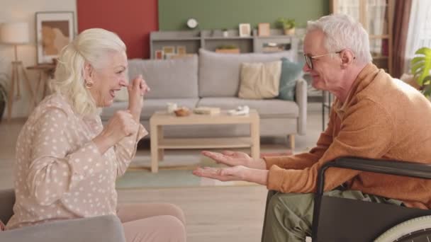 Widok z boku średni długi biały mężczyzna siedzi na wózku inwalidzkim przed swoją białowłosą żoną w salonie w ciągu dnia, para seniorów gra klapsy, śmieje się i rozmawia - Materiał filmowy, wideo