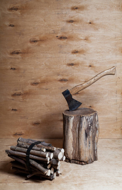 Ax and firewood. The ax sticks out in a stump against a background of light plywood. Nearby lies a bundle of round firewood. Stains are visible on a vertical plywood shee - Foto, Imagen