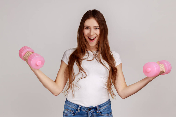Portrait of excited woman standing with raised arms and holding pink dumbbells, active lifestyle, wearing white casual style T-shirt. Indoor studio shot isolated on gray background. - Foto, Imagen