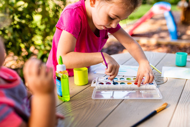 Child painting on paper outdoors on picnic table. Painting outside during the fall. Girl working on art project. - Zdjęcie, obraz