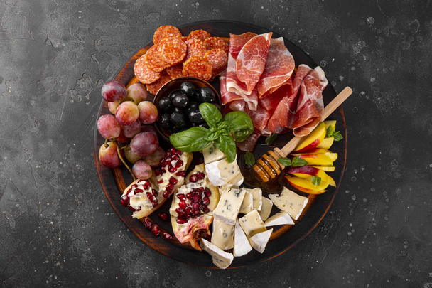  A set of appetizers for wine, jamon, pepperoni, cheese, grapes, peach and olives on a wooden board top view. Snack board on dark gray background. High quality photo - Foto, afbeelding