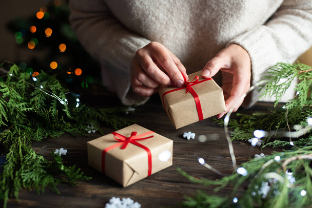 Γυναίκα περιτύλιγμα δώρο σε χαρτί χειροτεχνίας και κόκκινη κορδέλα, προετοιμασία δώρο για τα Χριστούγεννα, έννοια των διακοπών, close-up. - Φωτογραφία, εικόνα