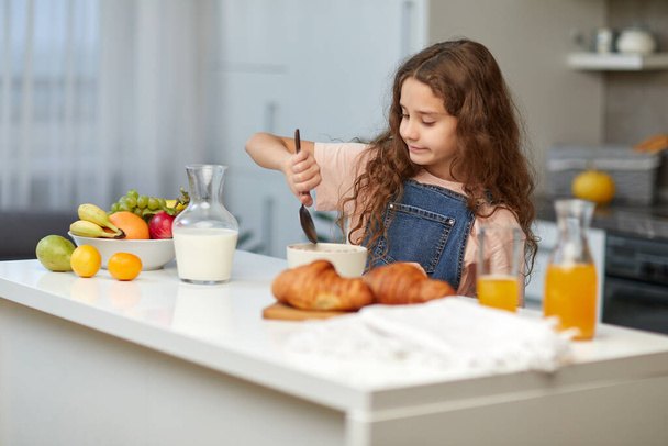Adorable niña con el pelo rizado comiendo cereales desayuno saludable en la cocina. - Foto, imagen