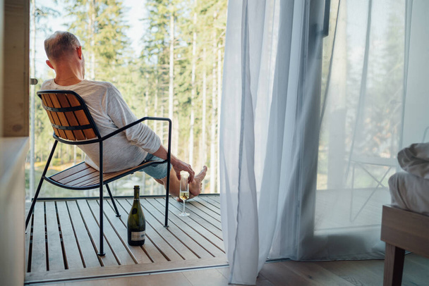 Sám muž sedí v křesle na balkóně venkovského domu a vychutnává si přírodní výhled na les, čerstvý vzduch a studené šampaňské víno sklenice. Pohled dozadu s otevřenou lahví na podlaze. Koncept oslav úspěchu. - Fotografie, Obrázek