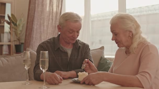 Chest-up radostné starší ženy krmení bělocha s narozeninovým dortem, sedí doma u stolu, s šumivým vínem, přičemž a usmívá - Záběry, video