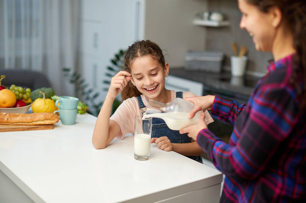 Portrait of a mom pours her cute laughing daughter milk in glass for breakfast - Foto, immagini