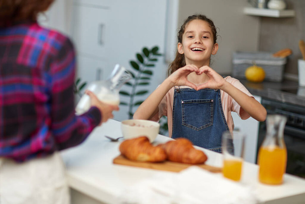Menina encaracolado bonito na cozinha com sua mãe, olhando para a câmera sorrindo fazendo forma de símbolo do coração com as mãos. - Foto, Imagem