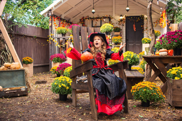 little girl dressed as a witch with a pumpkin in halloween autumn in the garden near the house - Фото, изображение