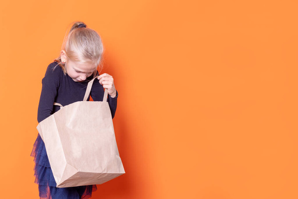 A beautiful Caucasian blonde girl in a black dress looks into an open craft paper bag on an orange background. - 写真・画像