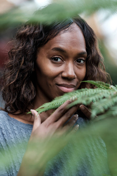 Снимок взрослой чёрной женщины, которая касается растения и смотрит в камеру.. - Фото, изображение