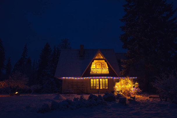 Linda casa de campo vintage decorada en el medio del bosque por la noche. Tranquila Nochebuena con nieve en el jardín y luz proveniente de la ventana. - Foto, Imagen