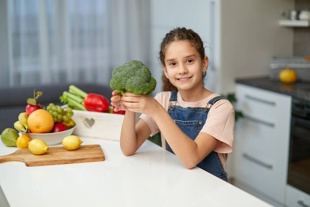 Adorable little curly girl wearing jeans and t shirt, holding broccoli, seated at table in the kitchen. - Photo, Image