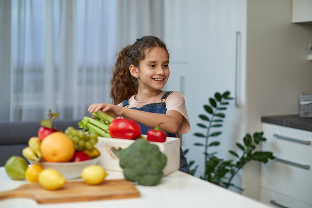 Adorable laughing and curly little girl sits at the table in the kitchen, near vegetables. - Photo, Image
