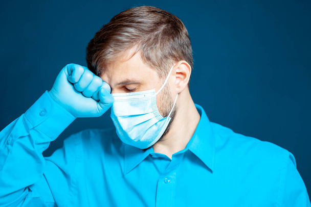 A doctor in a medical mask and gloves, in a blue uniform, leaned his fist against his head. - Photo, image