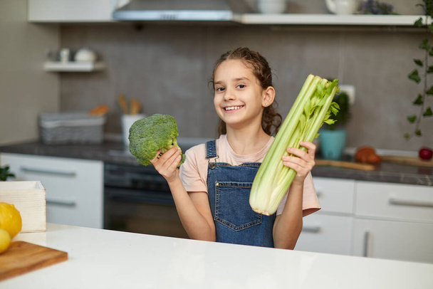 Nevető és göndör kislány áll az asztal mellett a konyhában, kezében zöldségekkel. - Fotó, kép