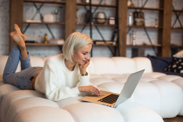 La giovane donna rilassata sul letto usa il computer portatile per lo shopping online e l'e-commerce con un sorriso gioioso. Femmina in accogliente camera da letto guardare video, navigare o scorrere i social media su pc portatile a casa al mattino - Foto, immagini